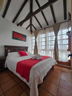 Une chambre à coucher avec une tête de lit en bois, un lit avec une literie blanche et rouge, un tableau représentant des fleurs accroché au mur et une fenêtre avec des rideaux.