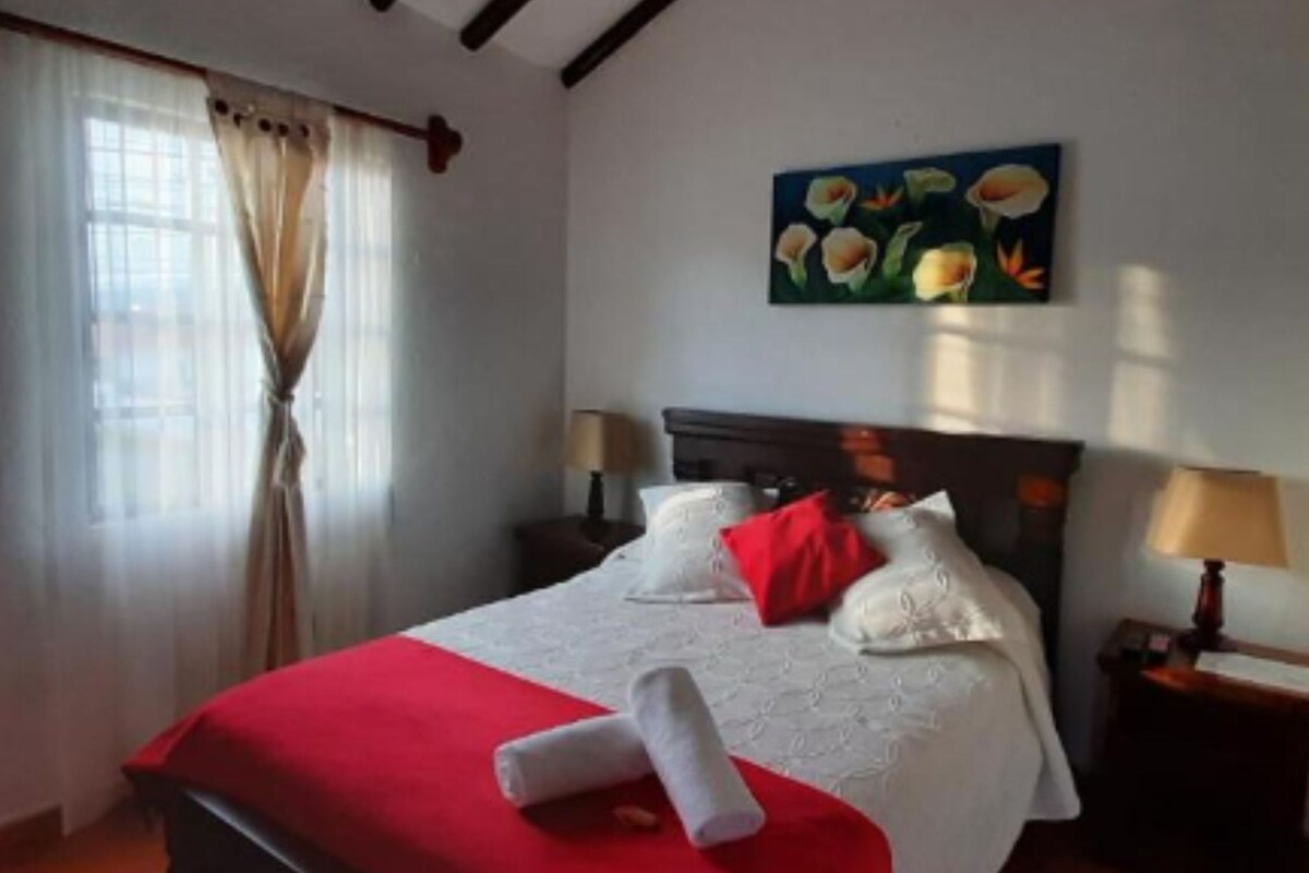 A hotel room with a bed, a painting of flowers, a window with curtains, and a nightstand with a lamp.