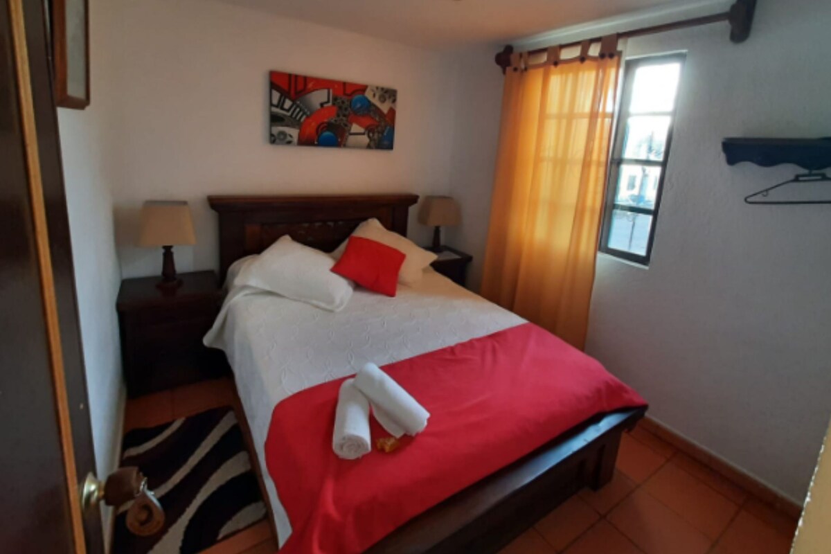 A hotel room with a bed, two bedside tables with lamps, a window with curtains, a wall-mounted shelf, and a painting above the bed.