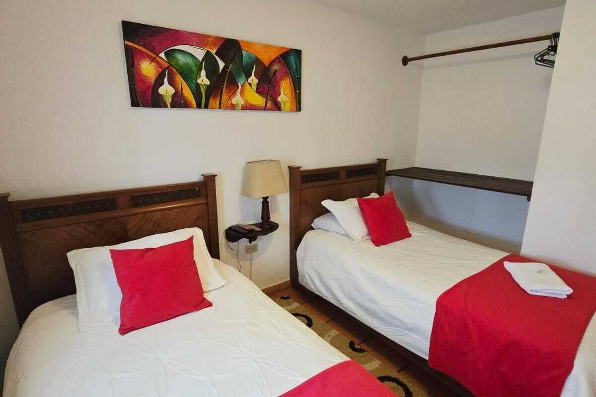 Habitación de hotel con dos camas, cada una con ropa blanca y almohadas rojas, una mesita de noche con lámpara, un cuadro en la pared y una luz en el techo.