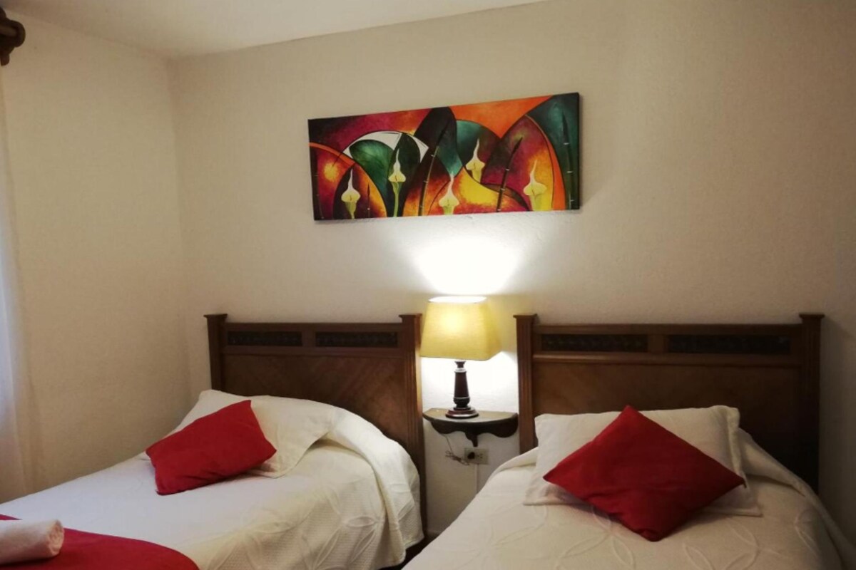 Une chambre d’hôtel avec deux lits, chacun agrémenté de draps et d’oreillers rouges, une table de chevet avec une lampe, et une peinture abstraite aux couleurs vives accrochée au mur.