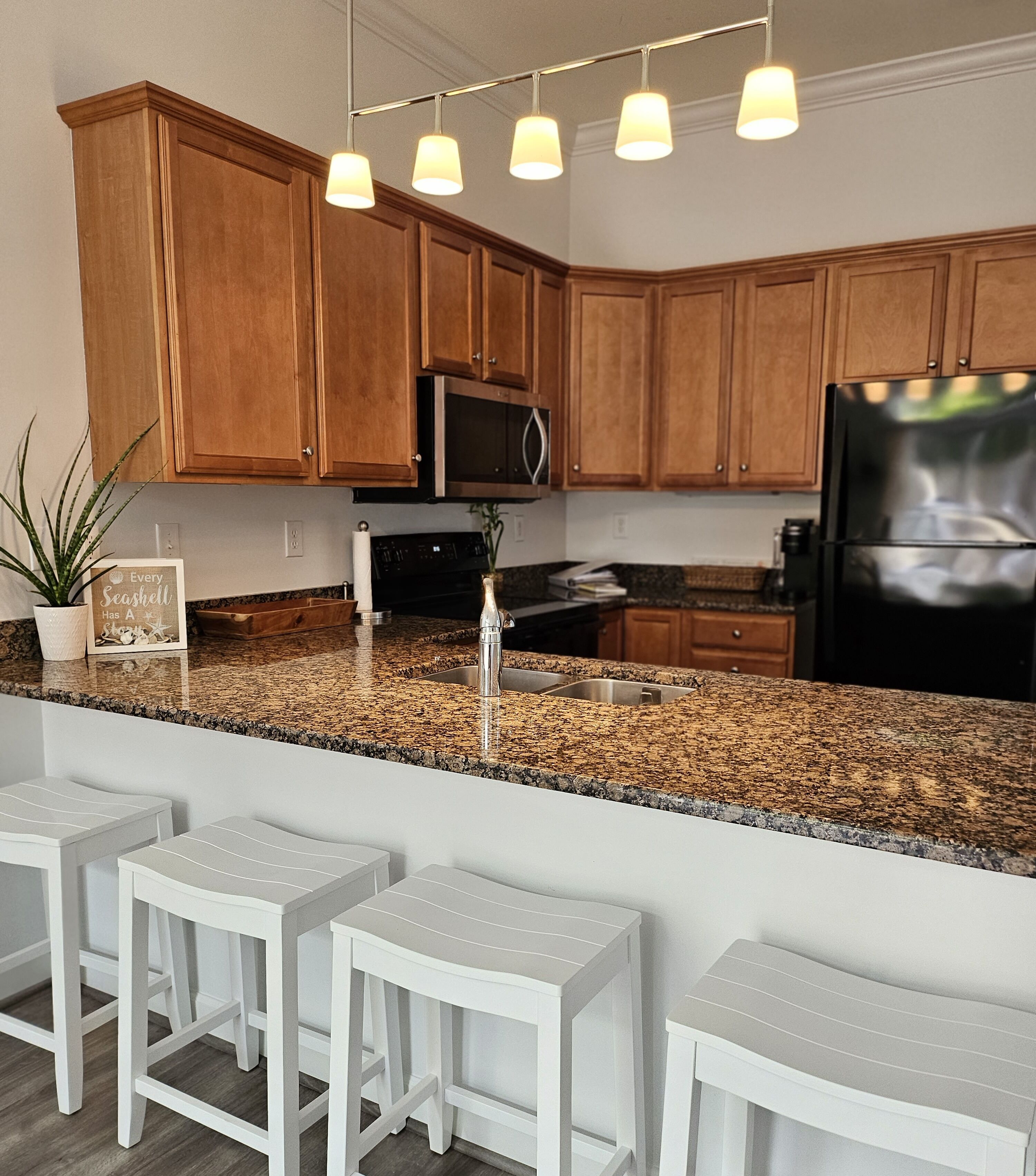 Una cocina con armarios de madera, encimera de granito, refrigerador de acero inoxidable y una barra con taburetes blancos.