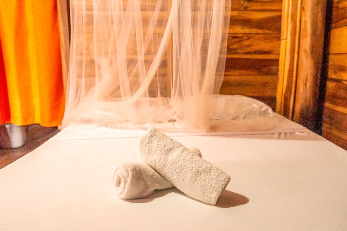 A bed with a white sheet and a rolled-up towel, a sheer white curtain, and a wooden wall.