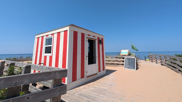 Un pequeño edificio blanco y rojo a rayas, con la puerta abierta, situado en un paseo de madera frente a la playa.