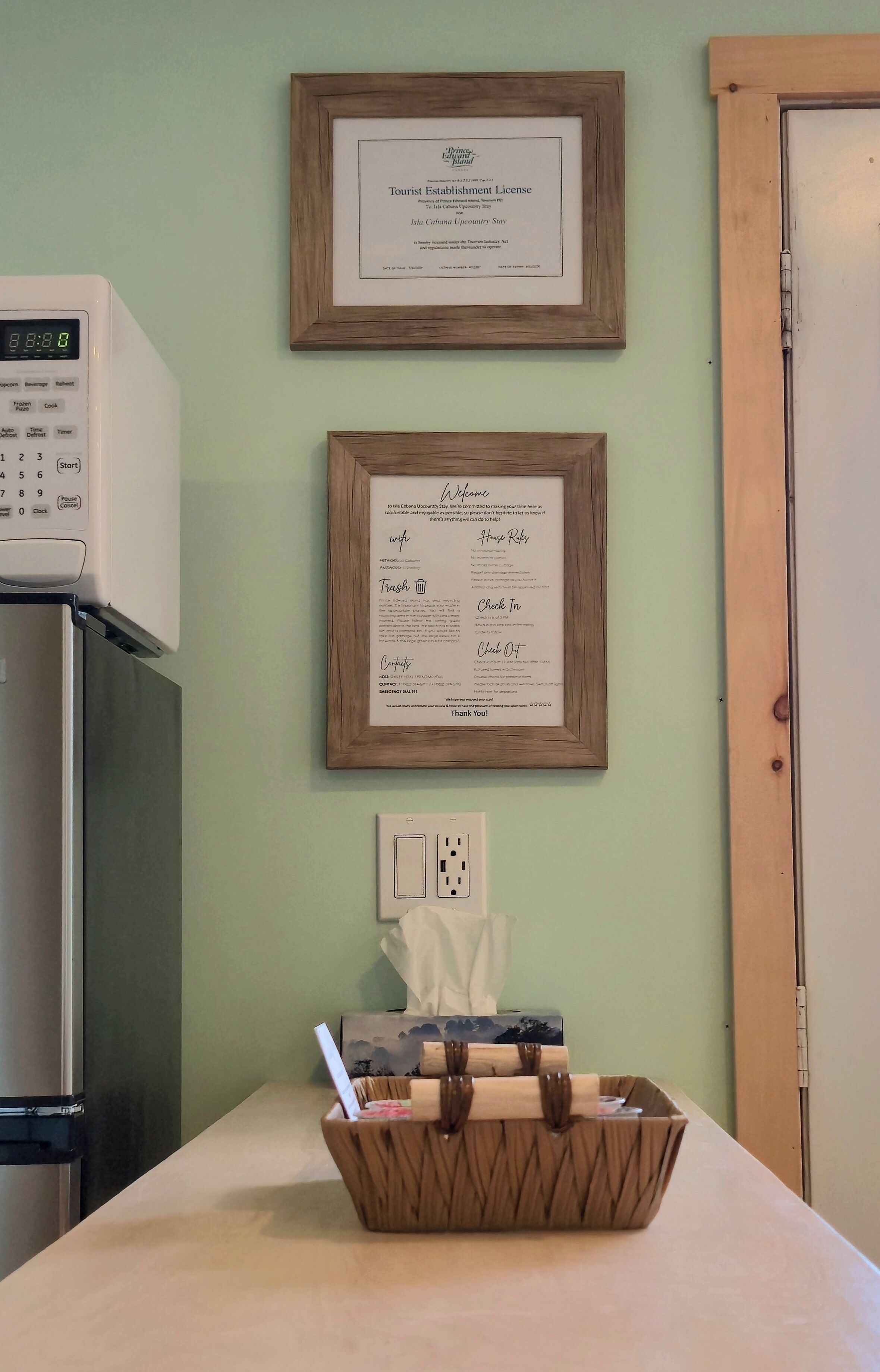 A kitchen with a microwave, a refrigerator, and a framed welcome sign on the wall.