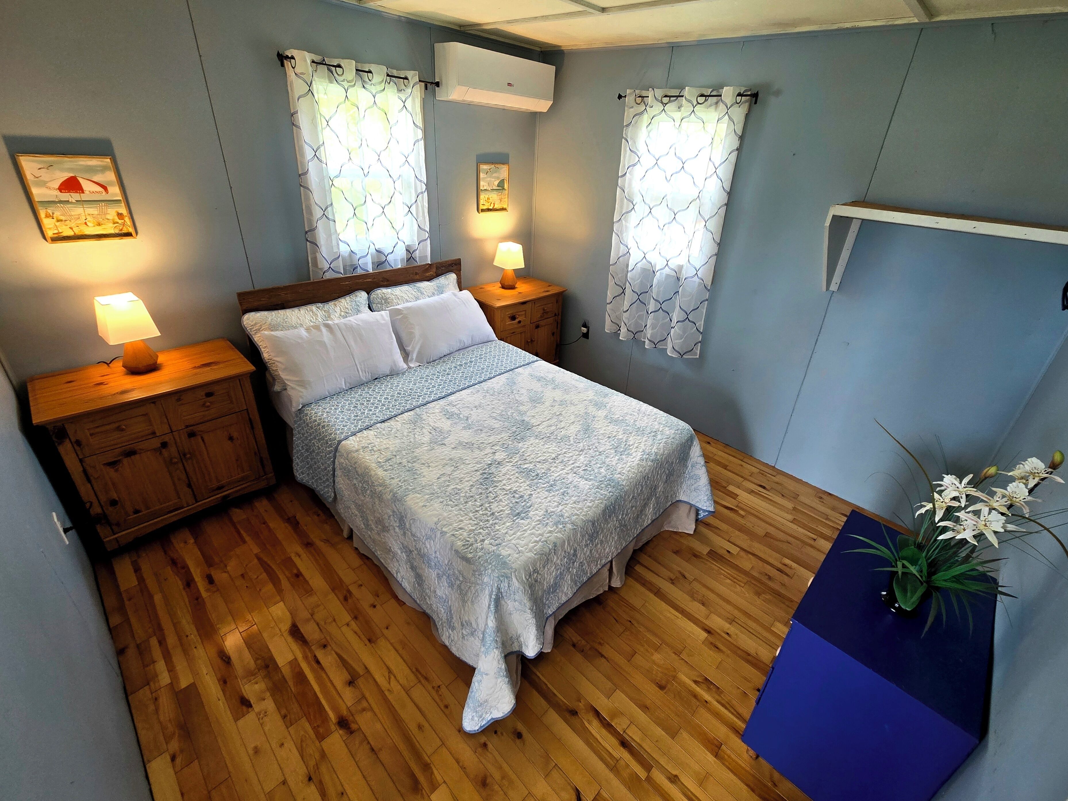 Une chambre à coucher avec un lit, des tables de chevet en bois, une lampe de chevet, un climatiseur mural et une plante en pot.
