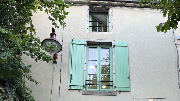Un edificio con una targa che riporta 'Rue du Pave', un muro in pietra con vegetazione, un tavolo di legno con sedie e una scala che conduce a un piano superiore.
