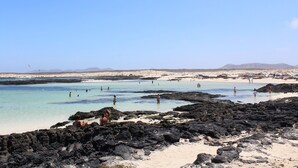 Une plage à l’eau claire, avec des zones rocheuses et des montagnes qui se dessinent à l’horizon.