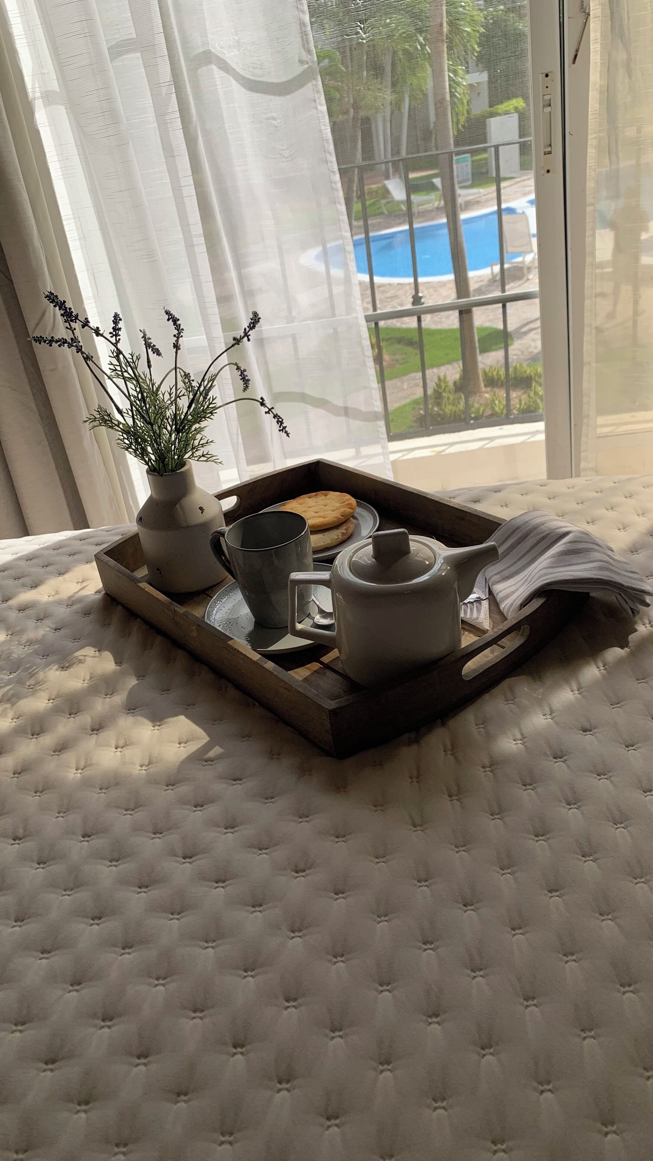 Uma bandeja com um bule, xícara e biscoitos sobre a cama, com vista para uma piscina.