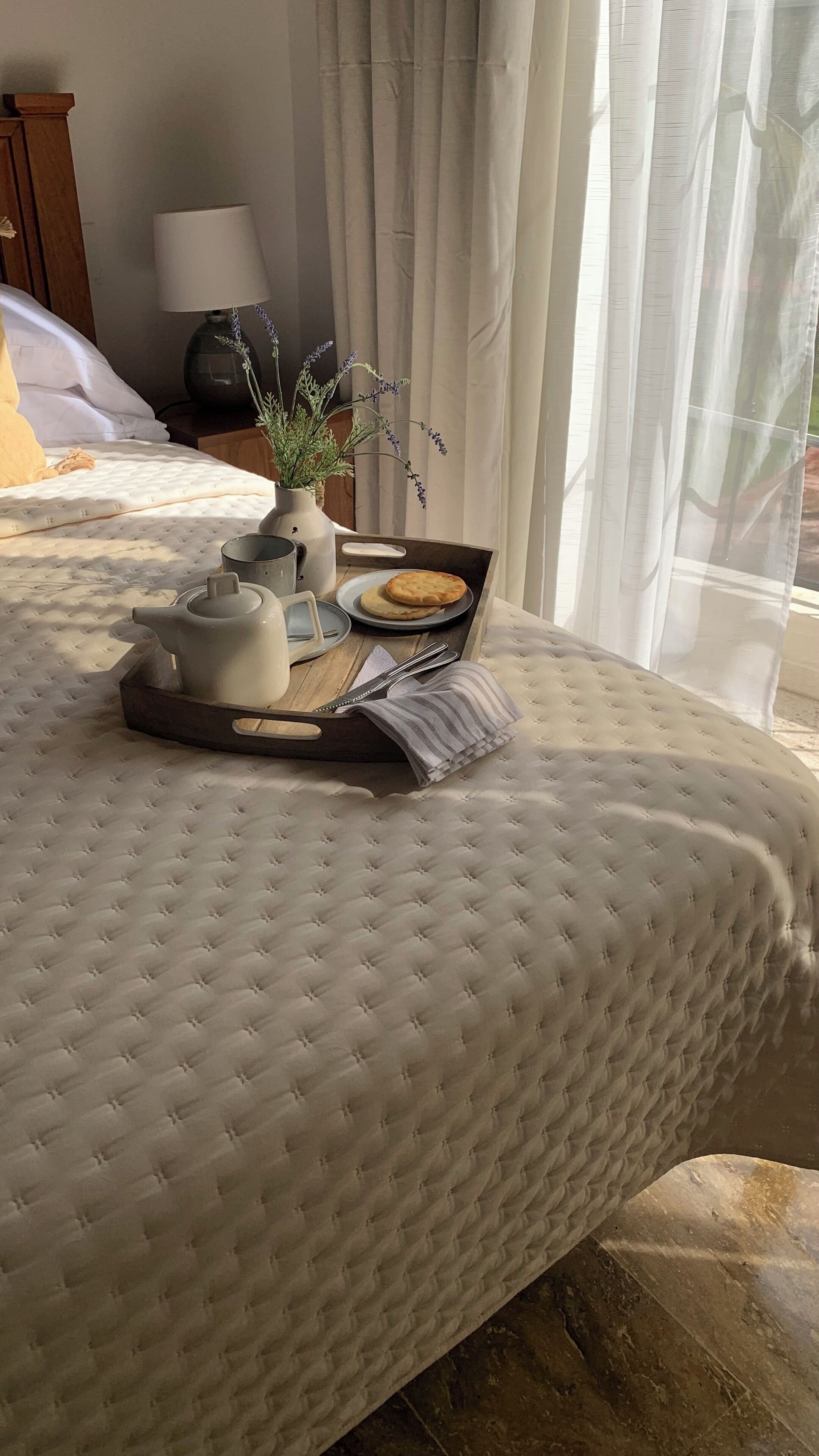 Un lit bien fait, avec une couvre-lit matelassé, un plateau avec une théière, une assiette de biscuits et un vase de lavande.