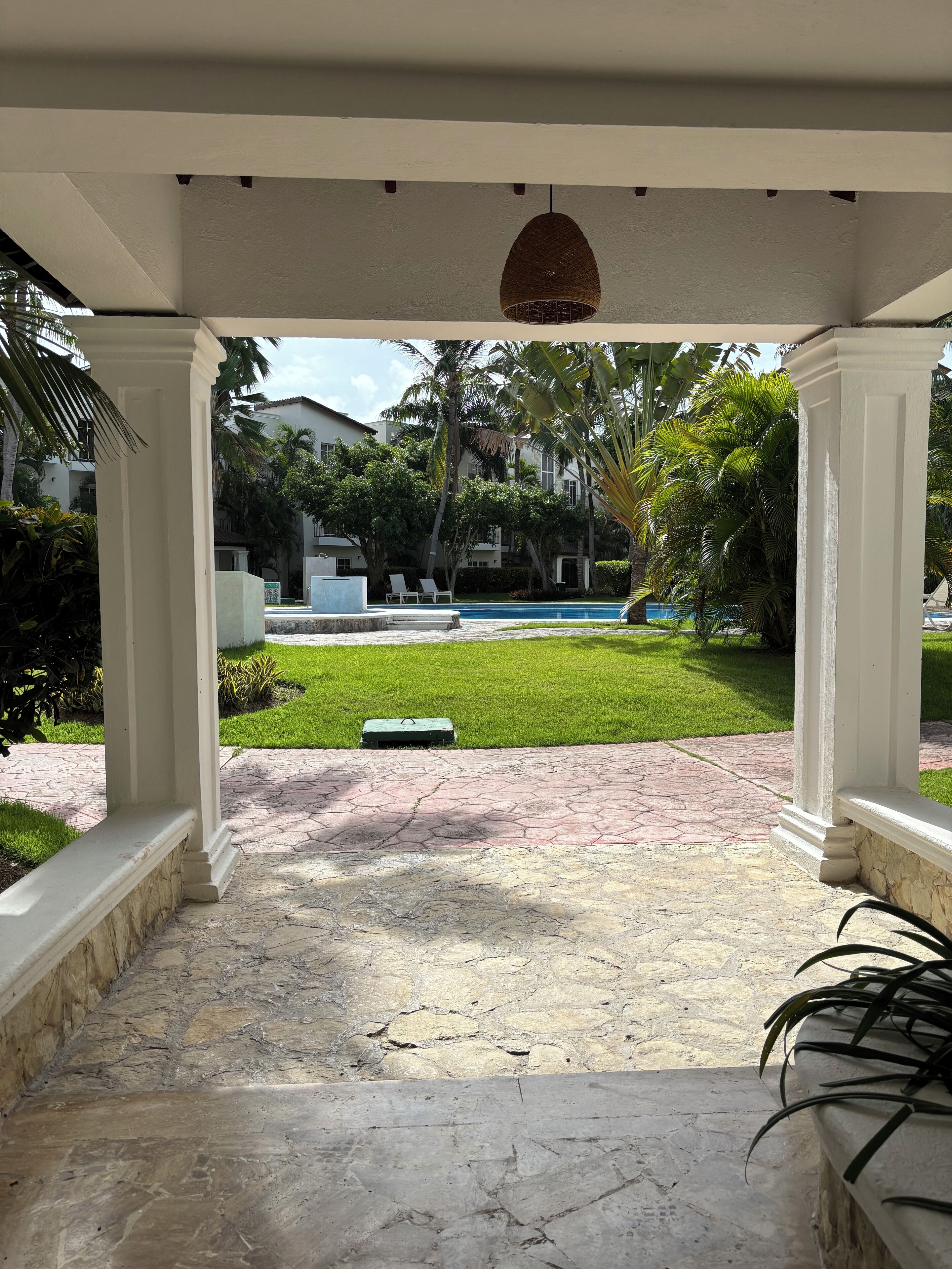 A covered patio area with a view of a swimming pool, palm trees, and a well-maintained lawn.