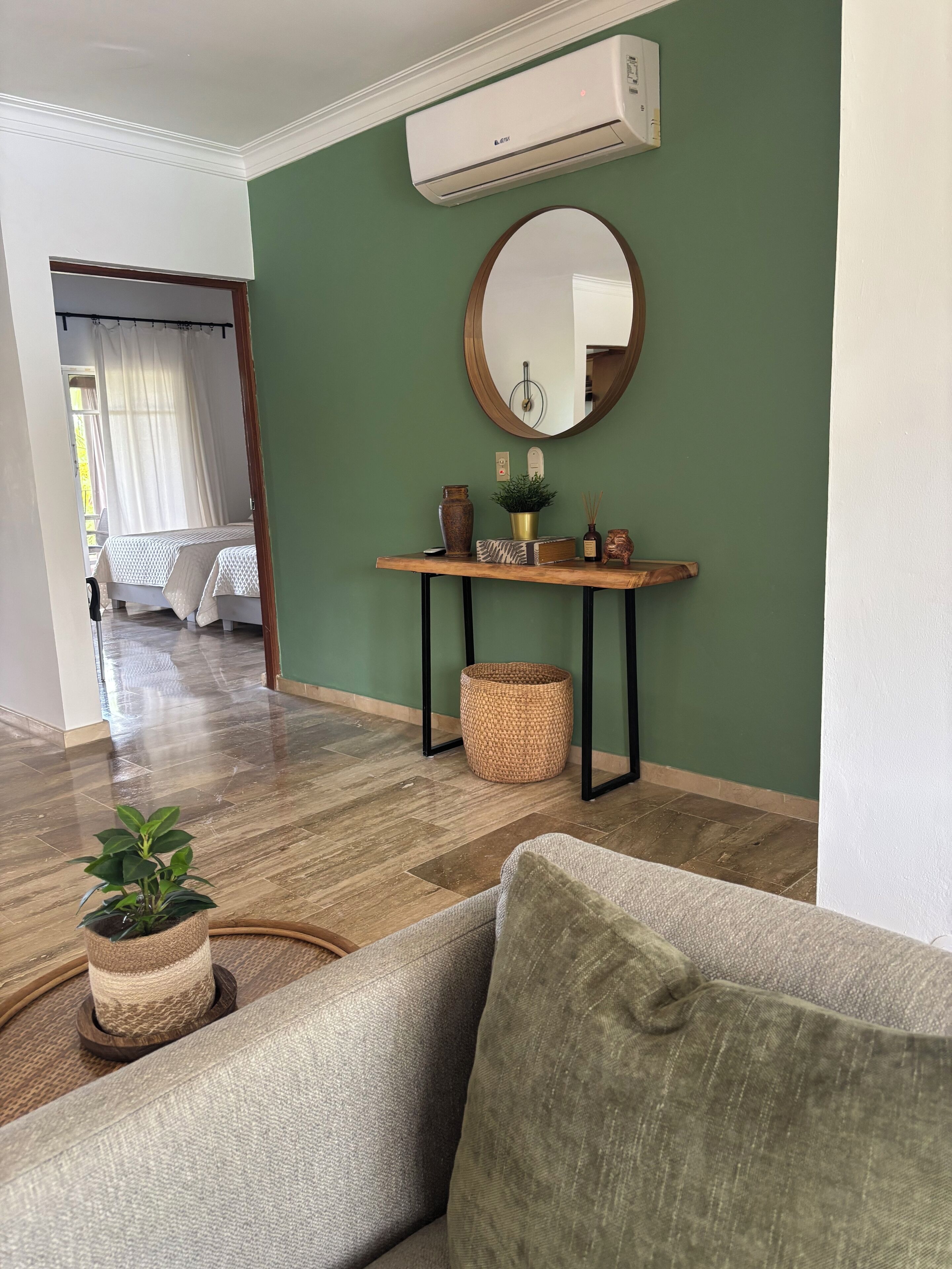 Un salón con pared verde, un espejo redondo, una consola de madera con adornos, un sofá gris y una cesta de mimbre.