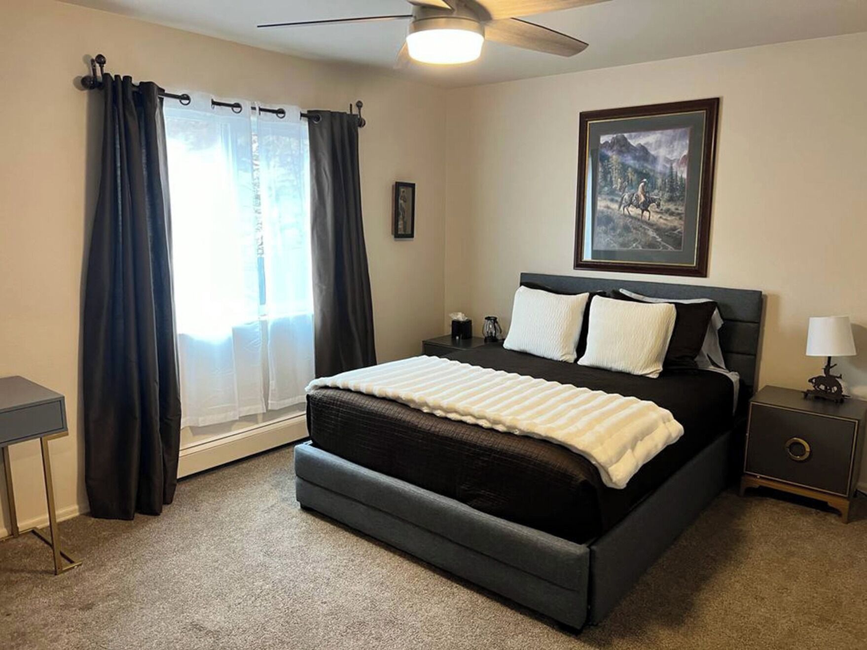 A bedroom with a bed, a nightstand, a lamp, a ceiling fan, and a window with curtains.