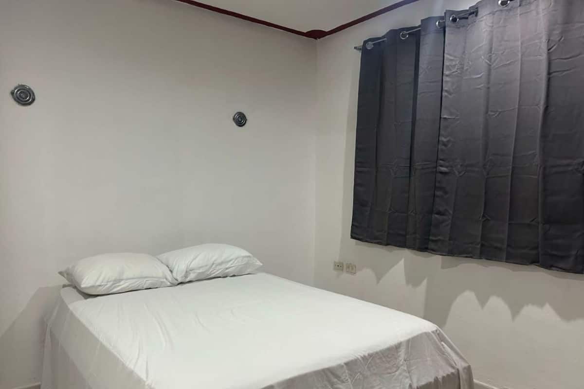 A bedroom with a bed, two pillows, a window with curtains, and a wall-mounted light fixture.