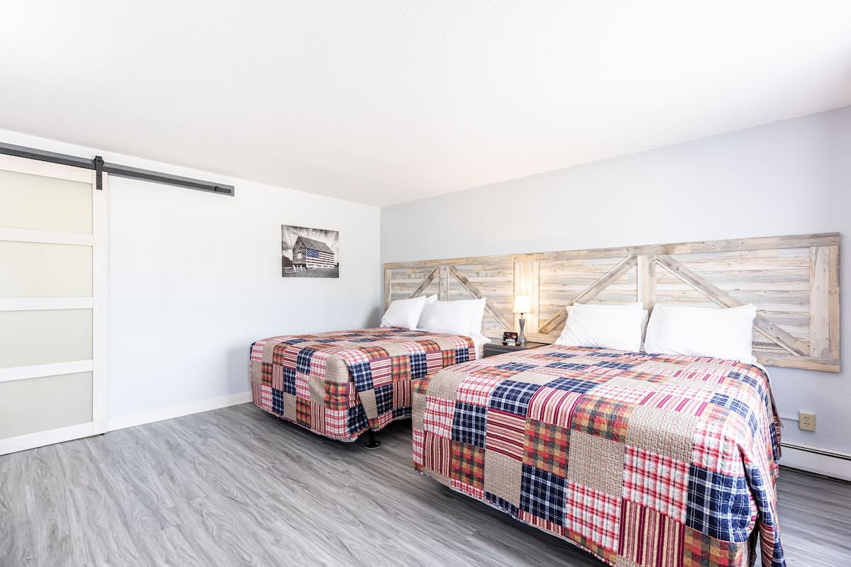 A room with two beds, each with a plaid bedspread, a wooden headboard, and a small bedside table with a lamp.
