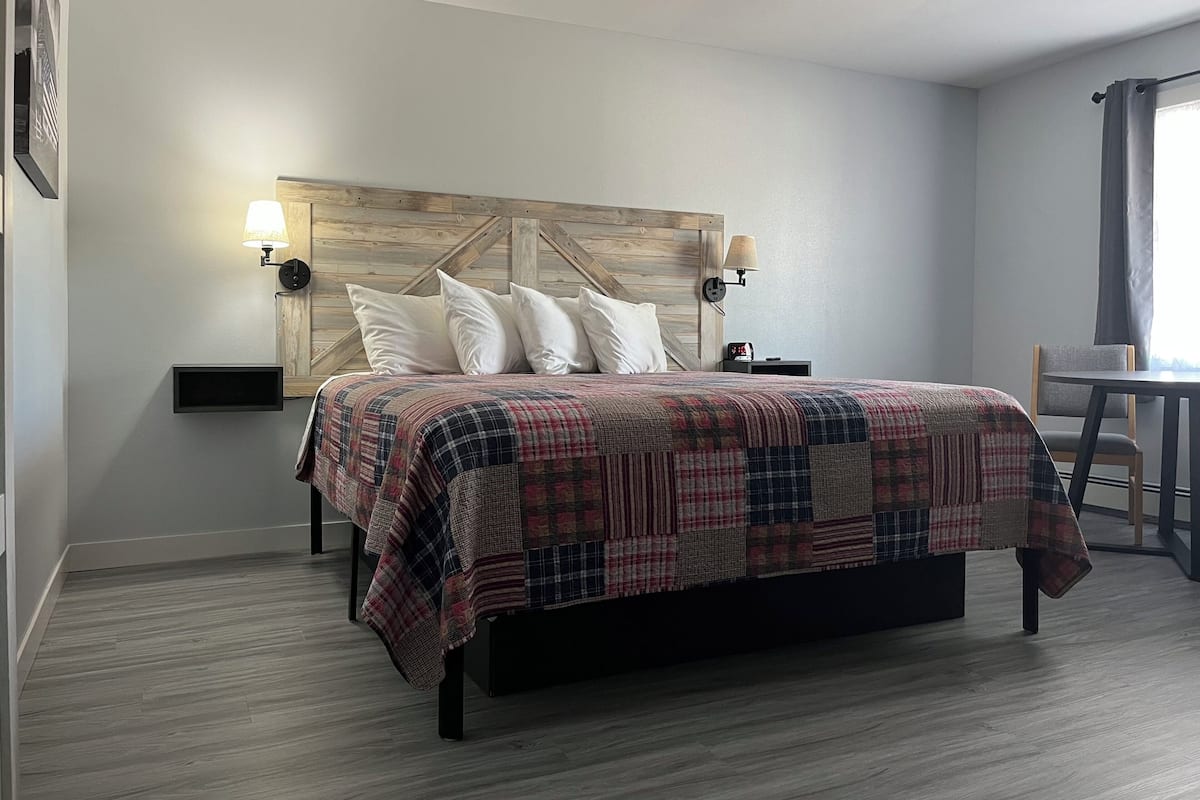 A neatly arranged bedroom with a wooden headboard, a plaid bedspread, two bedside lamps, and a small table with a chair.