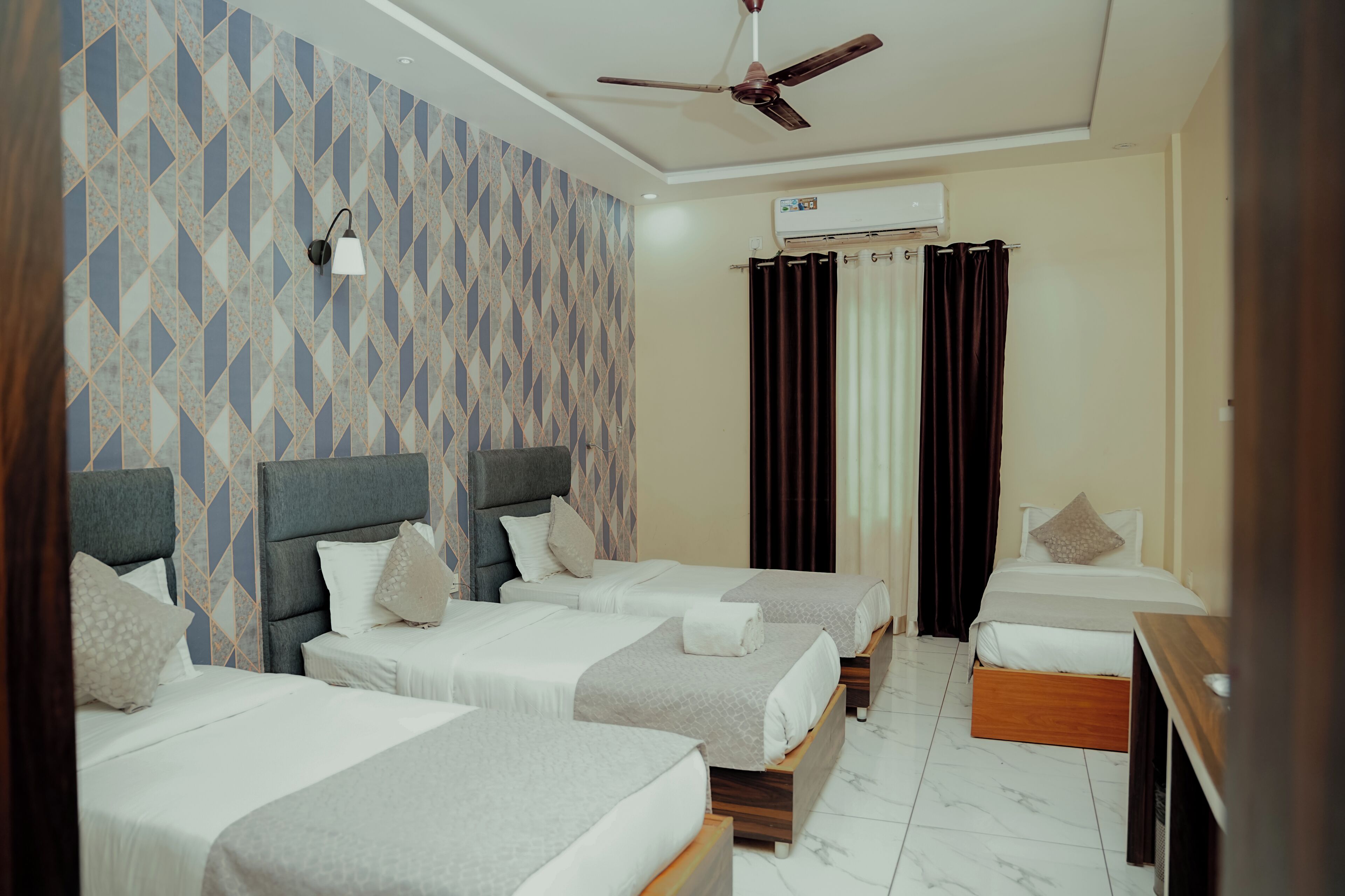 Une chambre d’hôtel avec trois lits, un ventilateur de plafond, un appareil de climatisation et un mur décoré de motifs.
