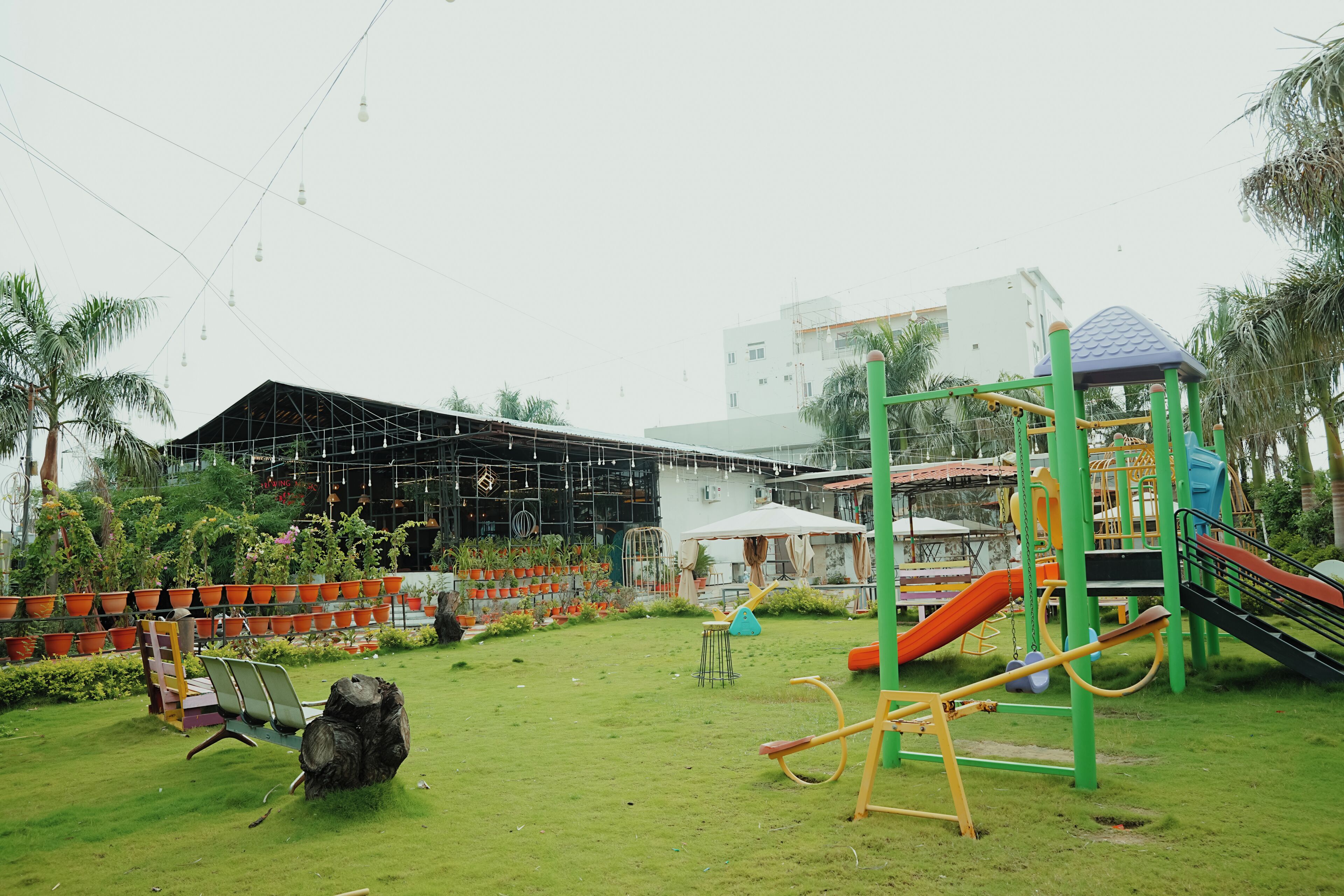 A playground with slides and swings, surrounded by potted plants and a building with a metal framework.