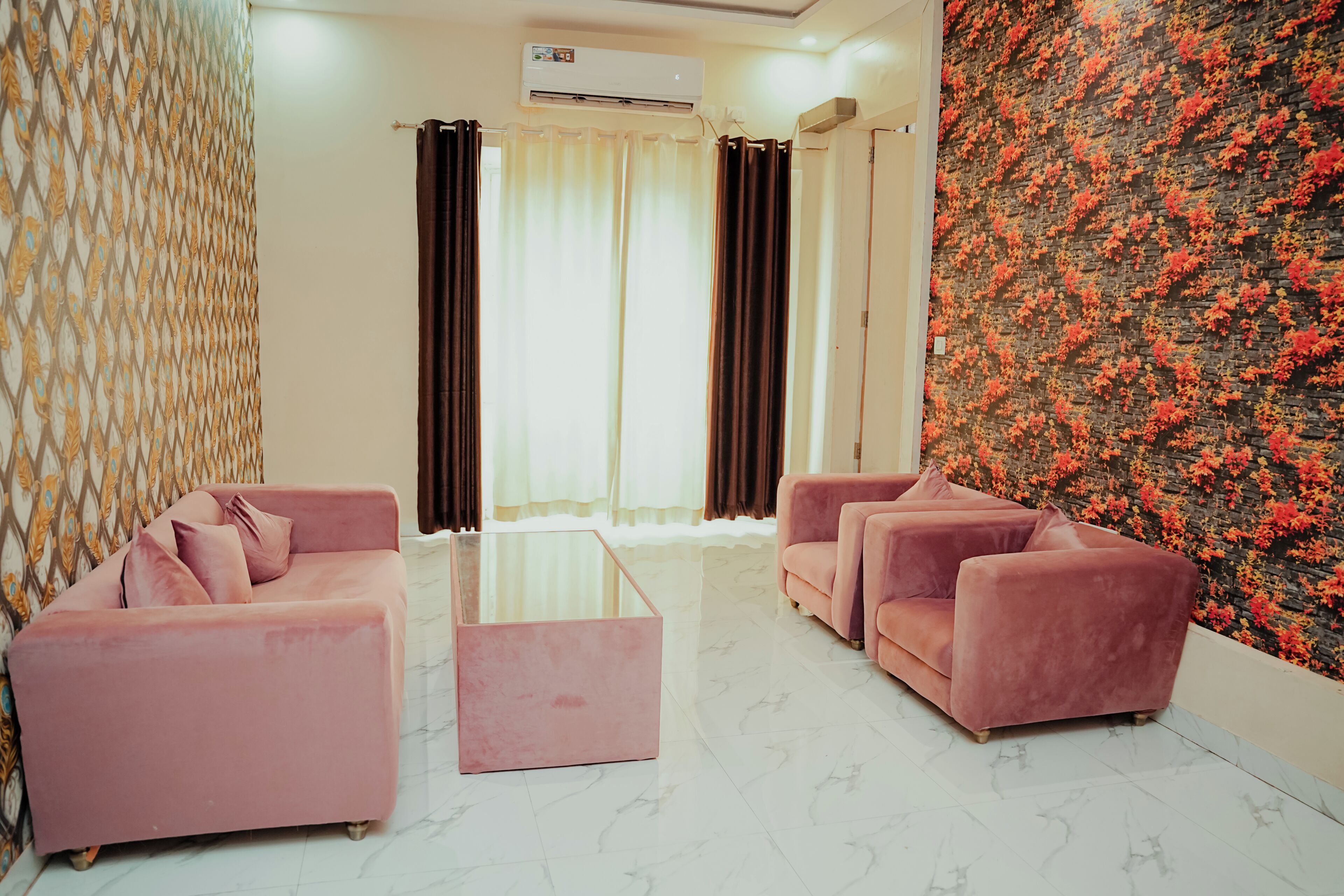 A modern living room with a glass coffee table, two pink sofas, a patterned wall, and a window with curtains.