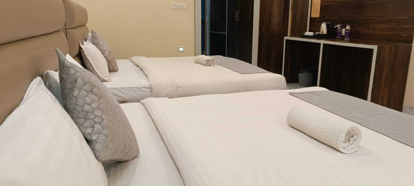 A hotel room with two beds, each with a pillow and a blanket. There is a small table with a kettle and cups.