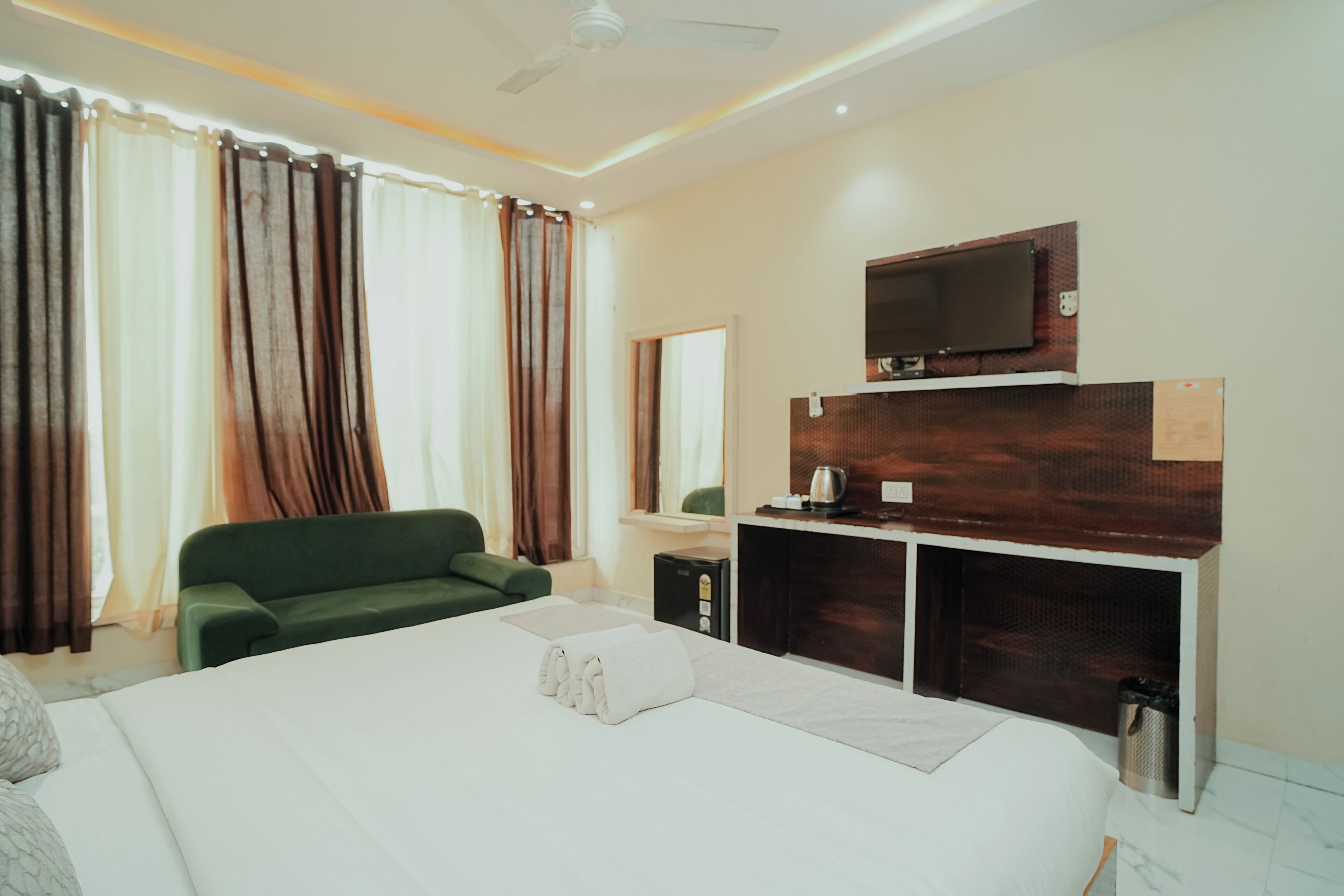 Habitación de hotel con cama, sofá verde, televisor montado en un panel de madera y una ventana con cortinas.