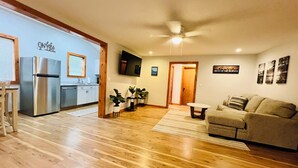 A spacious living area with a wooden floor, a sofa, a dining table, and a kitchen area with a refrigerator and a TV.
