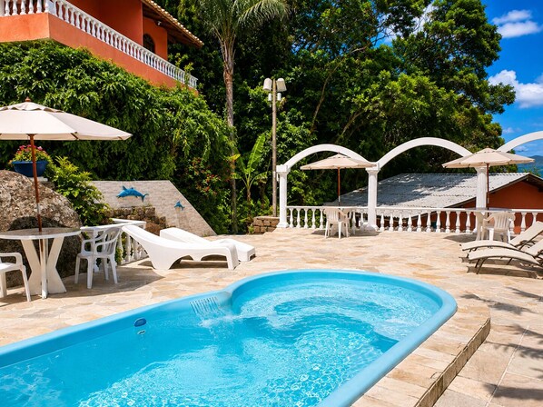 A swimming pool with lounge chairs and umbrellas, surrounded by a white fence and greenery.