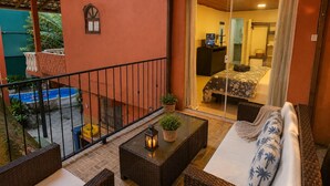 A balcony with wicker furniture, a small table with a lantern, and a view of a bedroom with a bed and a TV.
