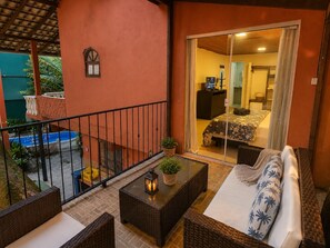 A balcony with wicker furniture, a small table with a lantern, and a view of a bedroom with a bed and a TV.