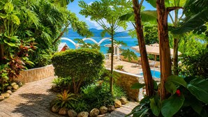 A lush garden with a pool, surrounded by tropical plants and a view of the sea.