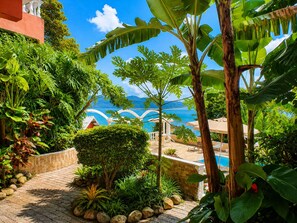 A lush garden with a pool, surrounded by tropical plants and a view of the sea.