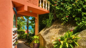 A pathway leading to a building with a balcony, surrounded by lush greenery and plants.