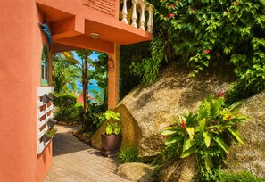 A pathway leading to a building with a balcony, surrounded by lush greenery and plants.
