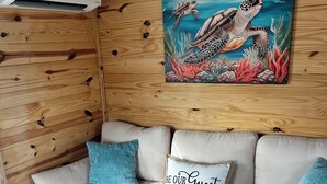 A cozy room with a wooden wall, a white sofa with blue cushions, a painting of sea turtles, and a sign that says "BE OUR GUEST".