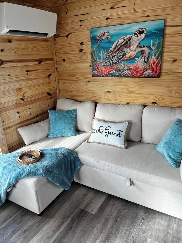 A cozy room with a wooden wall, a white sofa with blue cushions, a painting of sea turtles, and a sign that says "BE OUR GUEST".