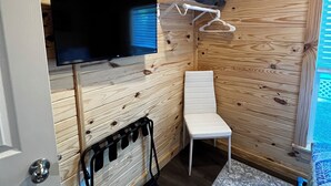 A compact room with a wooden interior, a mounted TV, a white chair, and a small shelf with a blanket.
