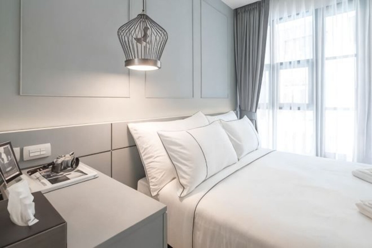A modern hotel room with a neatly made bed, a bedside table with a lamp, and a large window with curtains.