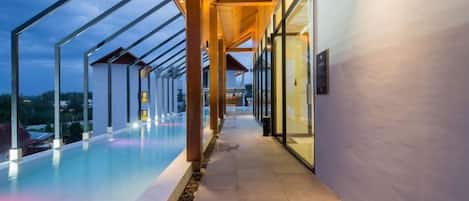 A modern building with a long corridor leading to a pool area, featuring glass walls and a wooden ceiling.
