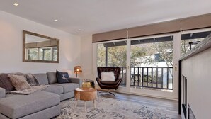A modern living room with a grey sectional sofa, a brown leather chair, a large window with a view, and built-in shelves with decorative items. - Ridge Creek 1 - Crackenback Ridge (Ski-In-Ski-Out)