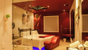 A modern hotel room with a glass partition, a bed, a red leather chair, and a small table with towels and a bottle. - K Hotel (León)
