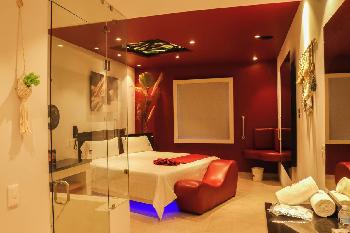 A modern hotel room with a glass partition, a bed, a red leather chair, and a small table with towels and a bottle.