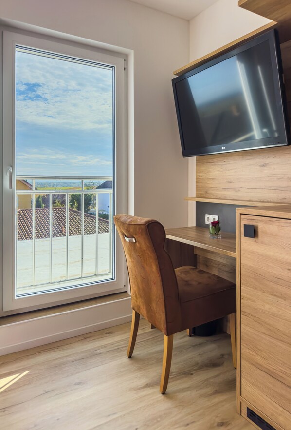 A modern room with a wooden desk, a brown leather chair, a flat-screen TV mounted above the desk, and a sliding glass door leading to an outdoor balcony with a tiled roof. - Hotel & Restaurant Traube (Kallstadt)