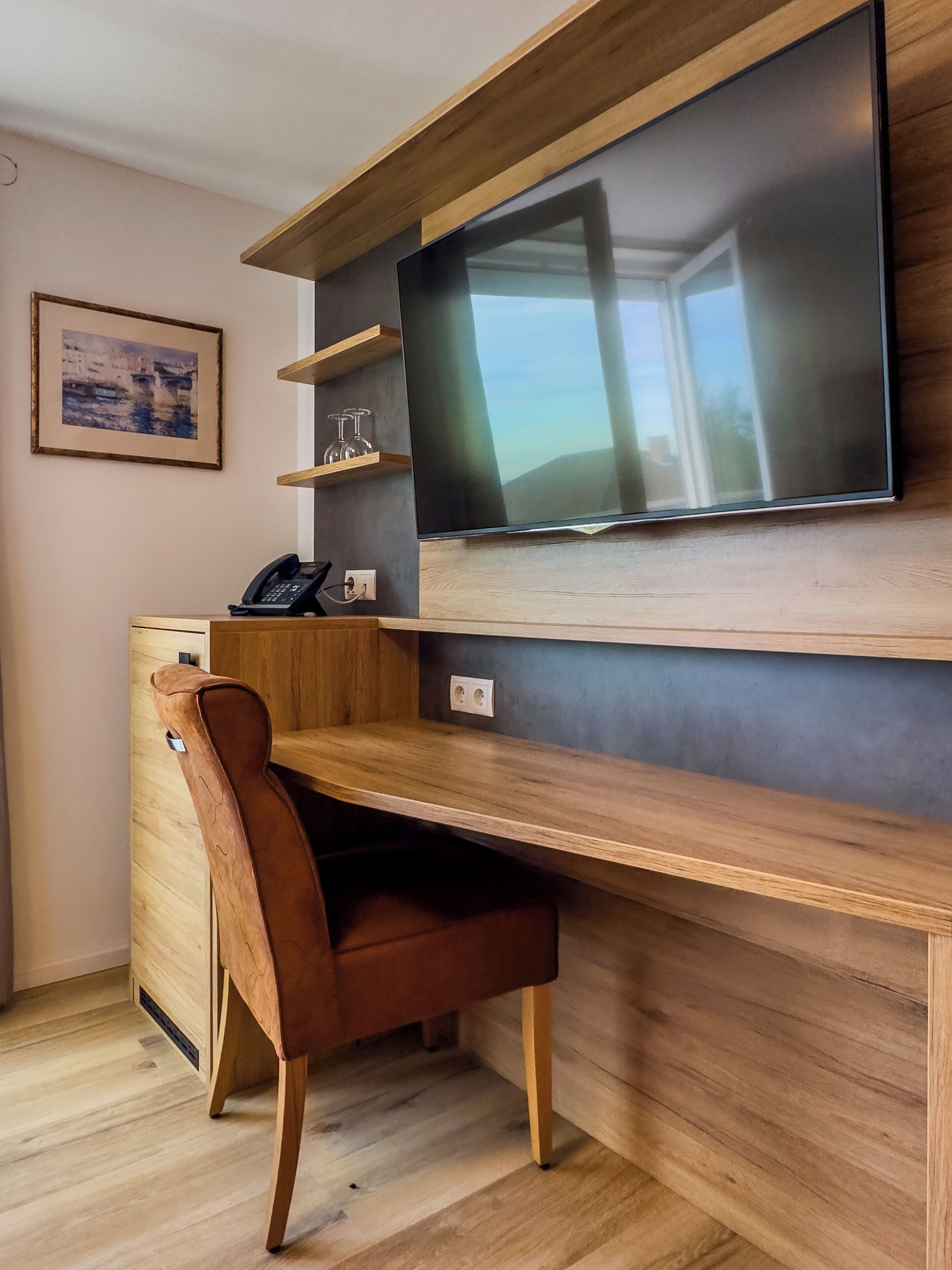Een moderne kamer met een houten bureau, een stoel, een flatscreen televisie aan de muur en een klein plankje met decoratieve objecten.