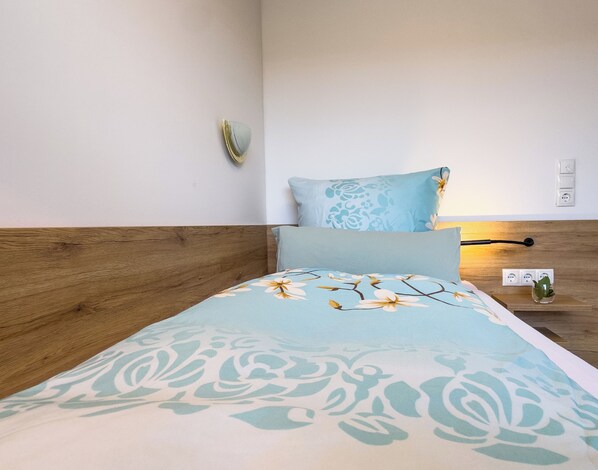 A neatly made bed with a floral bedspread, a wooden headboard, and a bedside table with a lamp and a small plant.