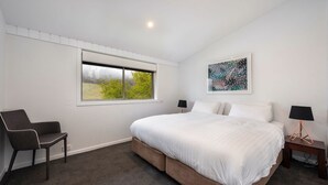 Un dormitorio con una cama grande, una silla, una mesita de noche, una lámpara, una ventana con vistas a la vegetación y un cuadro en la pared.