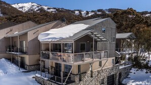 A modern multi-story building with balconies and a stone base, surrounded by snow and trees. - Snowbound Chalet 3 - Crackenback Ridge (Crackenback Ridge)