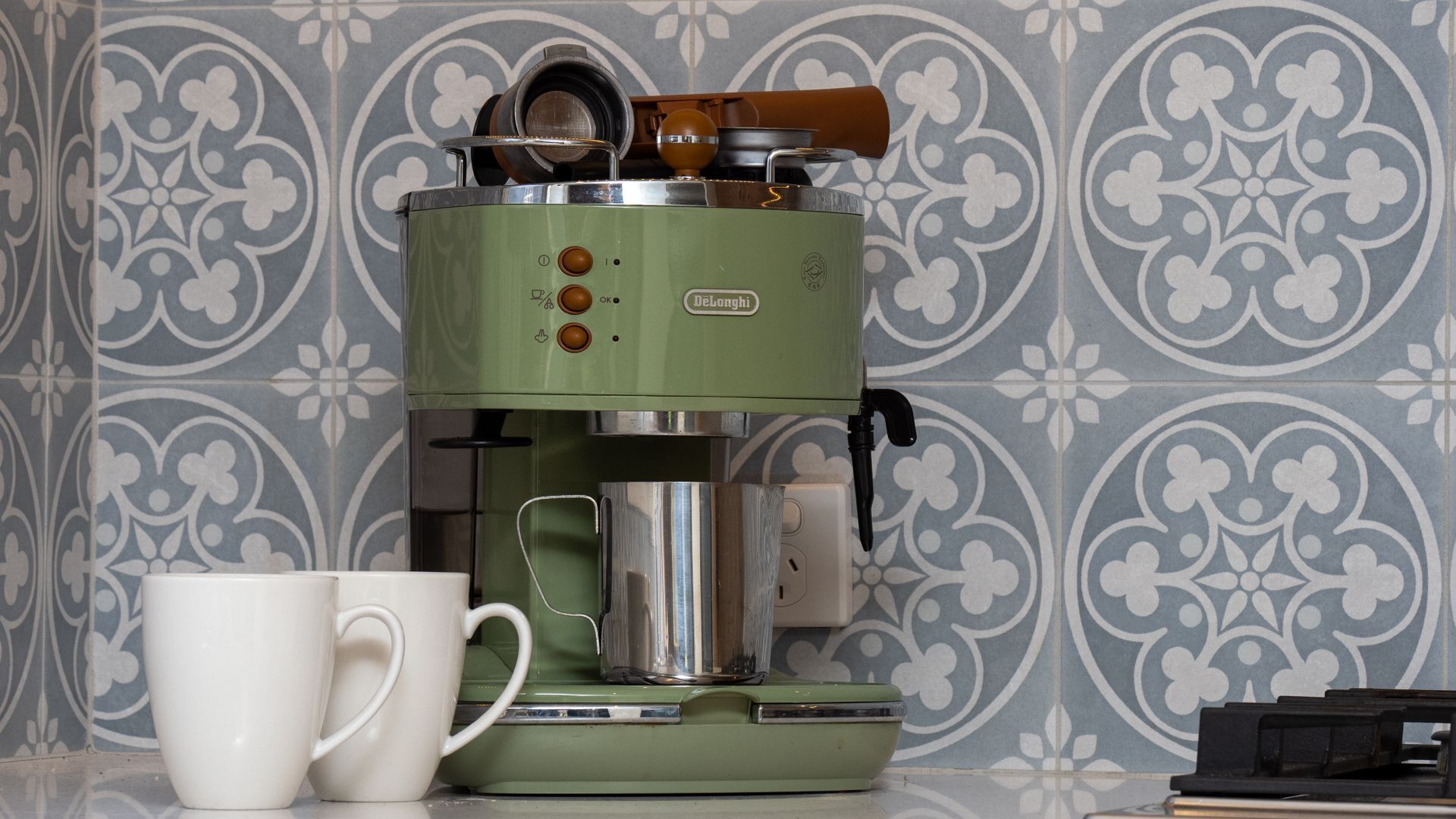 A vintage green espresso machine with two white cups beside it, set against a patterned tile backsplash.