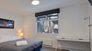 A bedroom with a bed, bedside lamps, a desk, a window with a view of a staircase, and a framed picture on the wall.