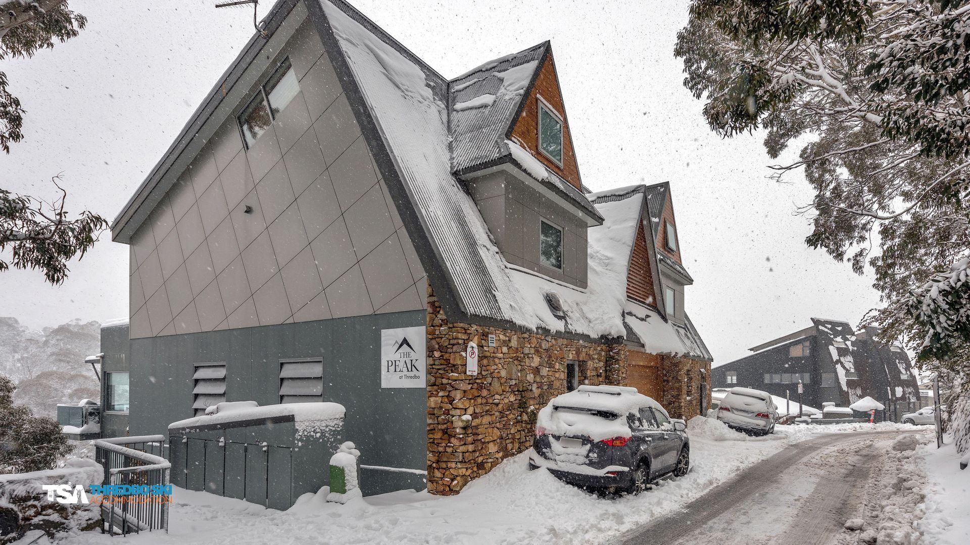 Un bâtiment à deux étages, doté d’une façade en pierre et d’un toit en pente, entouré d’arbres couverts de neige et de voitures garées.
