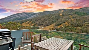 A balcony with a wooden table and chairs, overlooking a mountainous landscape with a sunset.
